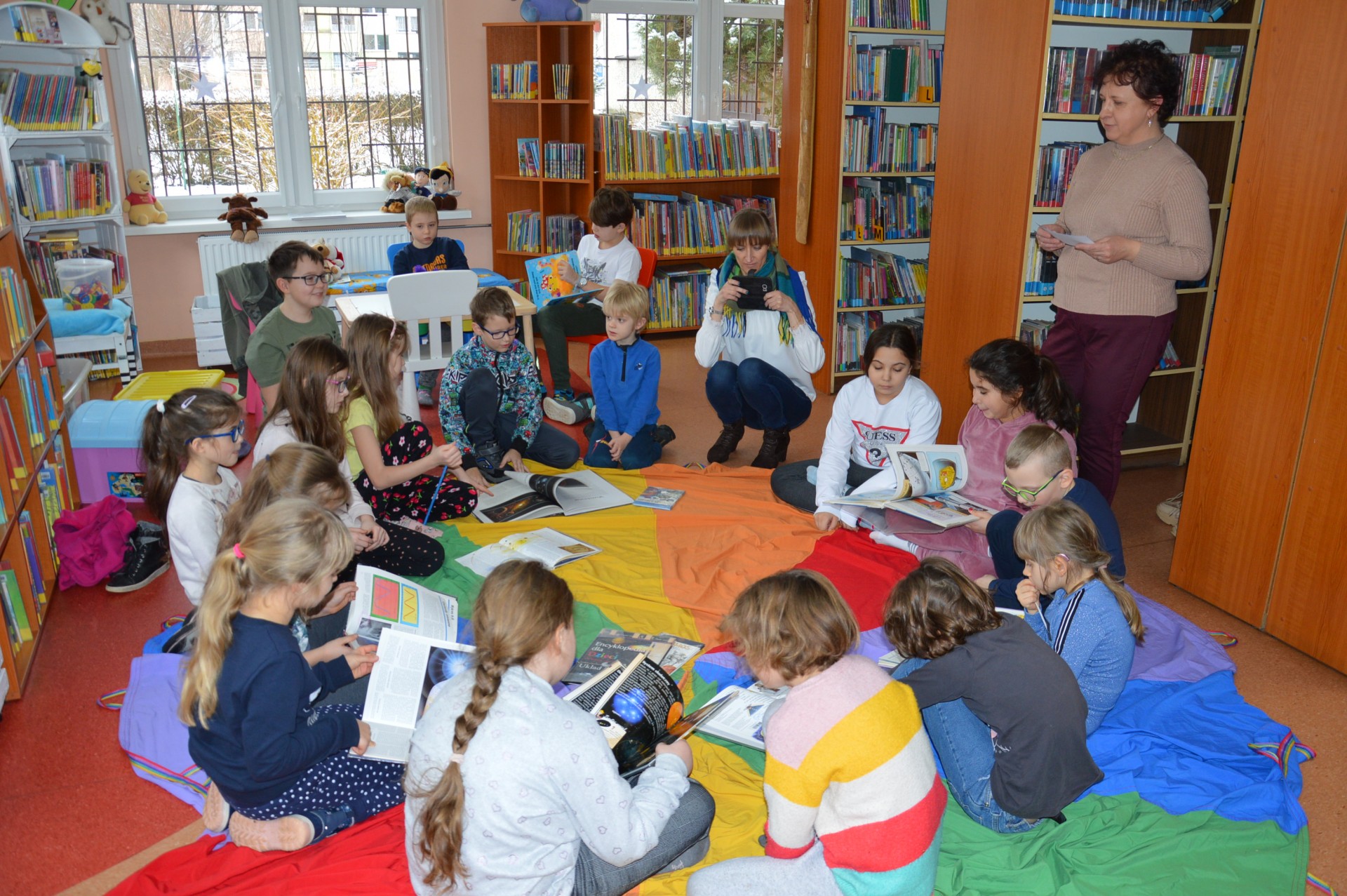 Dzieci siedzą wokół kolorowe okrągłej chusty. Czytają książki.